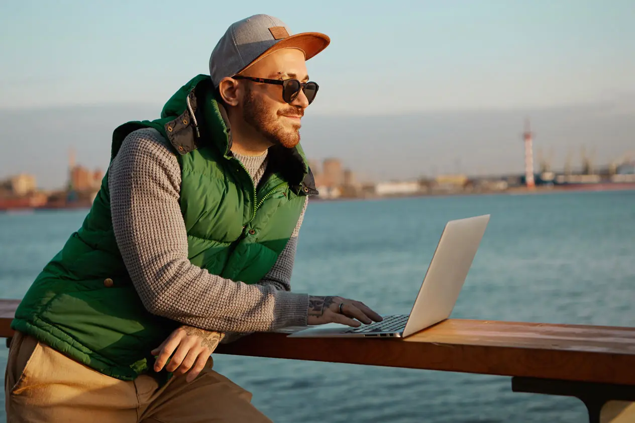 homme qui sourit en passant une commande en ligne sur son ordinateur posé sur une rambarde en bord de mer